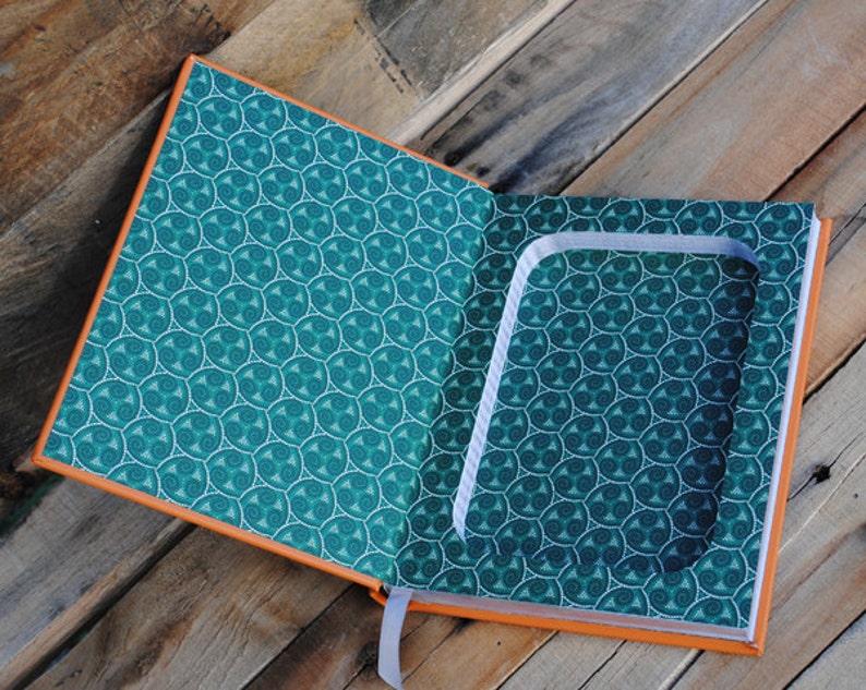 Puede incluir: Un libro abierto con un compartimento oculto en el interior. El libro tiene una cubierta de color turquesa con un patr&oacute;n de espiral y un borde naranja. El compartimento est&aacute; forrado con tela blanca.