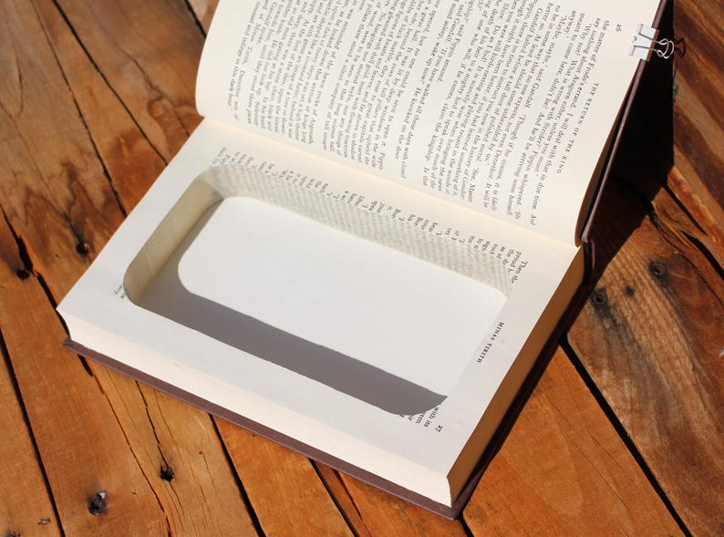 May include: An open book with a rectangular cavity cut into the pages, forming a hidden compartment. The book's pages are aged, and the edges are brown. The book is on a wooden surface.