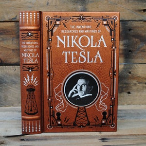 May include: A brown leather-bound book with a gold embossed cover featuring a portrait of Nikola Tesla and the text "THE INVENTIONS. RESEARCHES AND WRITINGS OF NIKOLA TESLA".