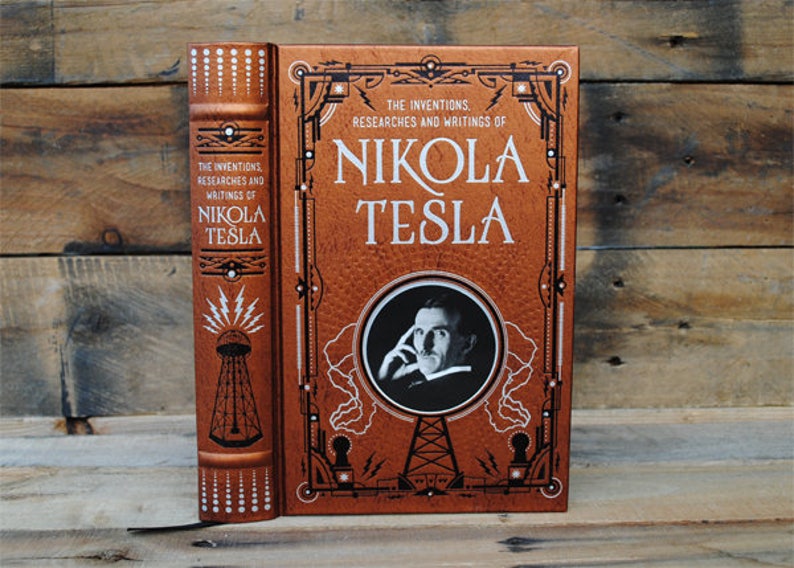 May include: A brown leather-bound book with a gold embossed cover featuring a portrait of Nikola Tesla and the text "THE INVENTIONS. RESEARCHES AND WRITINGS OF NIKOLA TESLA".