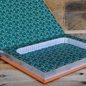 Puede incluir: Caja fuerte de libro abierta con un compartimento oculto. El libro tiene una cubierta con estampado verde y blanco y un exterior de cuero naranja. El compartimento est&aacute; forrado con un material blanco.