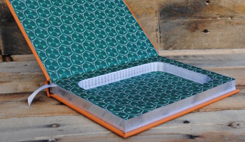 Puede incluir: Caja fuerte de libro abierta con un compartimento oculto. El libro tiene una cubierta con estampado verde y blanco y un exterior de cuero naranja. El compartimento est&aacute; forrado con un material blanco.
