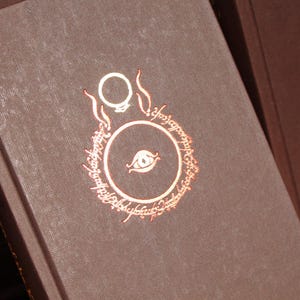 May include: Close-up of a brown book with a copper-coloured design featuring a ring and eye motif. The book's cover has a textured appearance. The design is surrounded by text. The book is part of a collection.