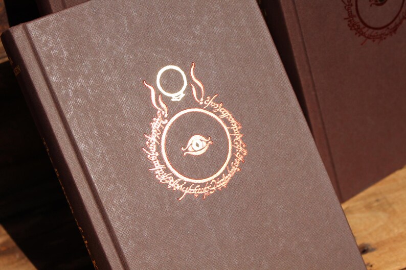 May include: Close-up of a brown book with a copper-coloured design featuring a ring and eye motif. The book's cover has a textured appearance. The design is surrounded by text. The book is part of a collection.