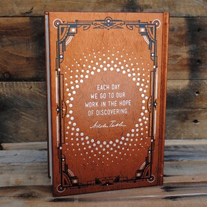 May include: A brown leather-bound book with a silver and black decorative design. The book features a quote: "Each day we go to our work in the hope of discovering." - Nikole Tech