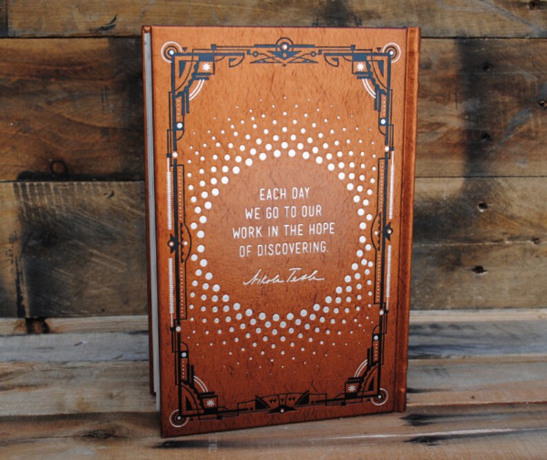 May include: A brown leather-bound book with a silver and black decorative design. The book features a quote: "Each day we go to our work in the hope of discovering." - Nikole Tech