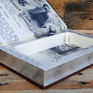 May include: A brown leather-bound book with a hidden compartment inside. The book is open, revealing a white foam lining and a black and white photo of a man standing in front of a large tower. The book's cover features the text "NIKOLA TESLA COMPANY" in blue.