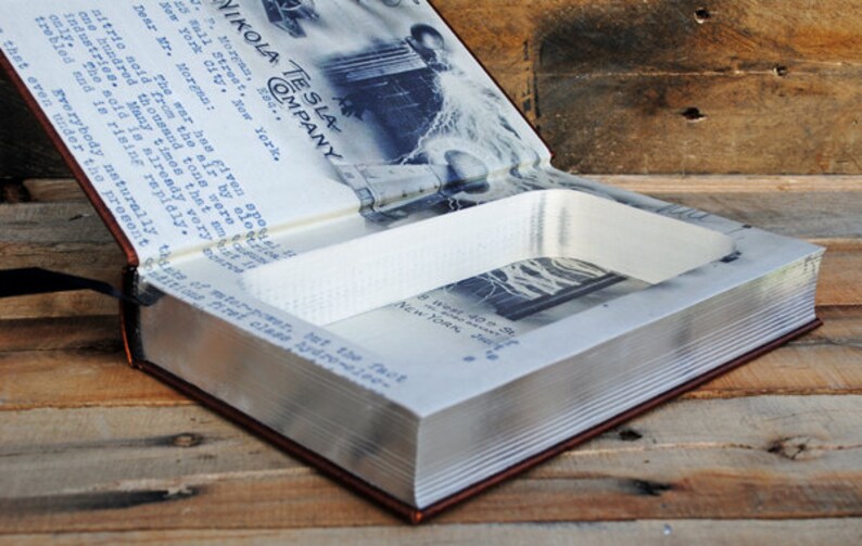 May include: A brown leather-bound book with a hidden compartment inside. The book is open, revealing a white foam lining and a black and white photo of a man standing in front of a large tower. The book's cover features the text "NIKOLA TESLA COMPANY" in blue.