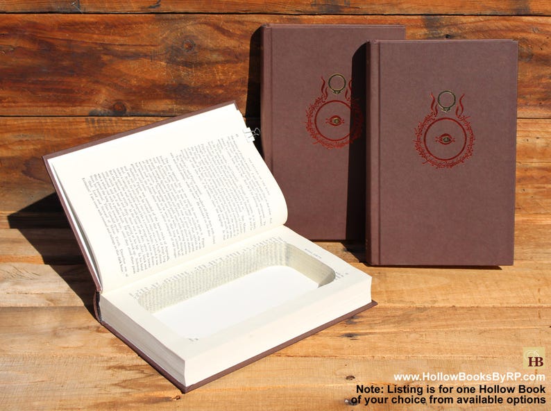 May include: A brown hollow book with a rectangular compartment, shown open. Two more books with brown covers and a red circular design are visible. The books are on a wooden surface. The website address www.HollowBooksByRP.com is visible.