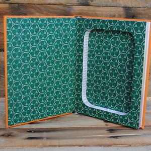 Puede incluir: Un libro naranja con una cubierta de patr&oacute;n verde est&aacute; abierto para revelar un compartimento oculto. El compartimento est&aacute; forrado con papel blanco y tiene un fondo de patr&oacute;n verde.
