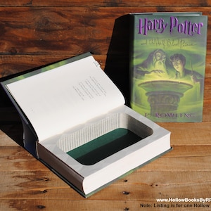 Può includere: Un libro svuotato intitolato "Harry Potter e il principe mezzosangue". Il libro è aperto, rivelando una cavità rettangolare con un interno verde. La copertina è verde e viola. Appoggiato su una superficie di legno.