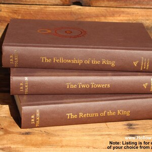 May include: Three brown hardback books stacked on a wooden surface. The titles are "The Fellowship of the Ring", "The Two Towers", and "The Return of the King" in gold lettering. The author's name, J.R.R. Tolkien, is also visible.