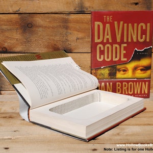 May include: A hollowed-out book with a rectangular cavity carved into the pages. The book is open, revealing the interior, with a red cover and the title "The Da Vinci Code" visible. The book is on a wooden surface.