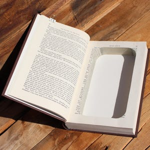 May include: An open book with a rectangular cavity cut into the pages. The book has a brown cover and rests on a wooden surface. A metal clip secures the pages.