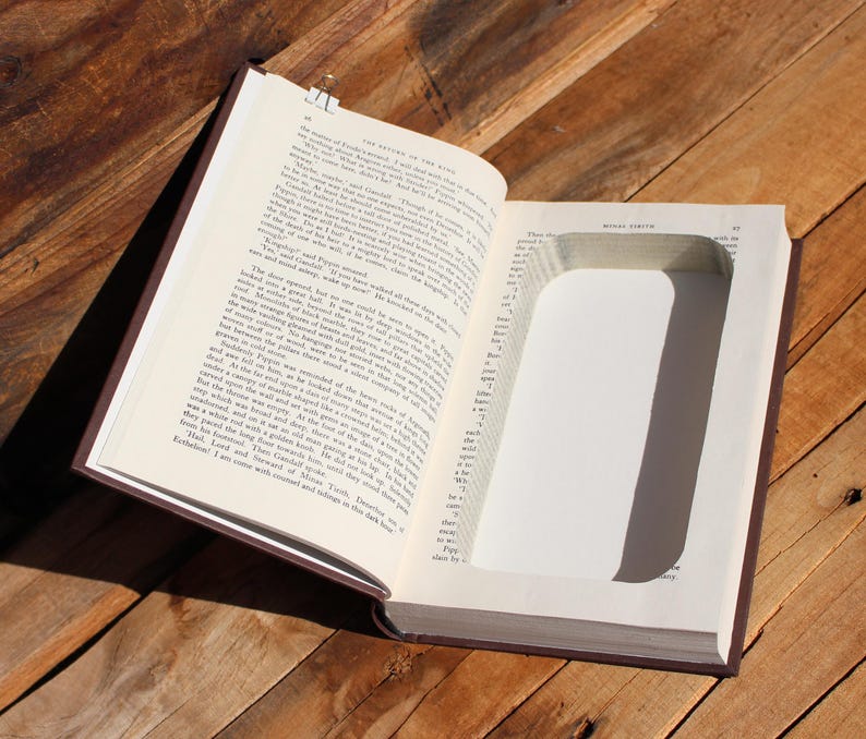 May include: An open book with a rectangular cavity cut into the pages. The book has a brown cover and rests on a wooden surface. A metal clip secures the pages.