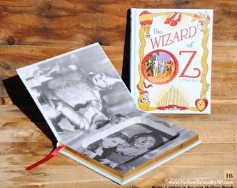 Puede incluir: Un libro azul claro con un compartimento oculto. La portada del libro presenta una ilustración de los personajes de El Mago de Oz y el texto "The Wizard of Oz" y "L. Frank Baum". El libro está abierto para revelar una foto en blanco y negro del Hombre de Hojalata y Dorothy de la película.