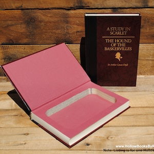 May include: A hollow book titled "A Study in Scarlet" and "The Hound of the Baskervilles" by Sir Arthur Conan Doyle. The book is open, revealing a pink interior with a rectangular cutout. The cover is dark red with gold lettering.