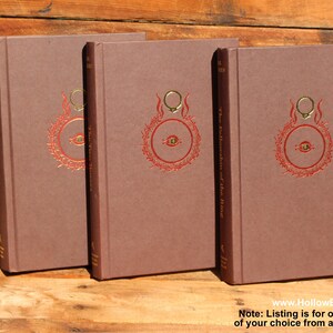 May include: Three brown hardback books with a red and gold design on the cover. The design includes a ring and an eye. The text "The Fellowship of the Ring" is visible on the spine. The books are on a wooden surface.
