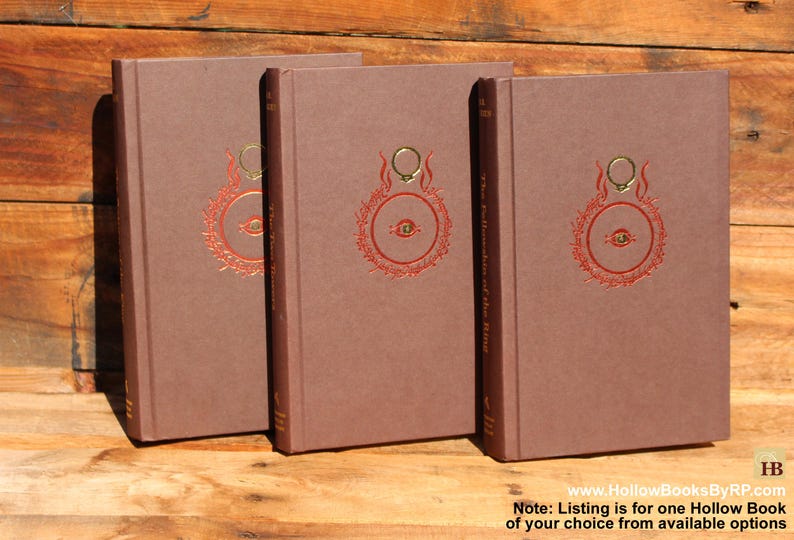 May include: Three brown hardback books with a red and gold design on the cover. The design includes a ring and an eye. The text "The Fellowship of the Ring" is visible on the spine. The books are on a wooden surface.