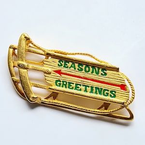 May include: A gold-colored sled-shaped brooch with the words "SEASONS GREETINGS" in green lettering on a wooden plank design. A red arrow points left. The brooch has a rope detail.