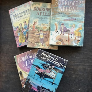 May include: A collection of vintage children's books, including titles like "The Borrowers Afloat" and "Bedknob and Broomstick." The book covers feature colorful illustrations and the authors' names. The books are arranged on a dark wooden surface.