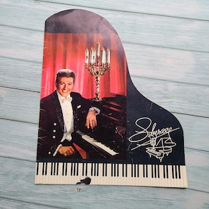 May include: A black and white image of a man in a tuxedo playing a grand piano. The piano has a black and white keyboard and a black finish. The man is wearing a white bow tie and a white shirt. The background is a red curtain with a candelabra in the center. The image is a vintage style advertisement for a piano.
