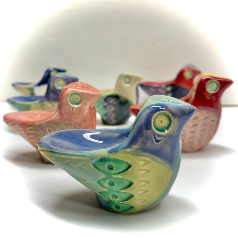 May include: A collection of six ceramic bird figurines, each with a unique colour scheme and intricate details. The birds are arranged in a row, with the blue bird in the foreground. The birds have a glazed finish and are hand-painted with various colours, including blue, green, pink, and red.
