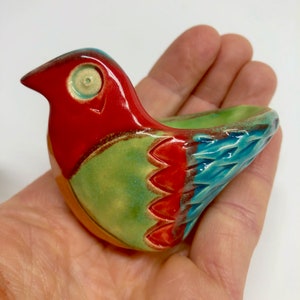 May include: A ceramic bird-shaped dish with a red, green and blue glaze. The dish has a small, round opening on the top and a larger, oval opening on the bottom.