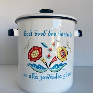 May include: A white enamel canister with a lid, featuring two side handles and a dark blue rim. The front displays a colorful floral design in red, yellow, and blue, with the Swedish text "Eget bord den bästa är av alla jordiska gåvor".