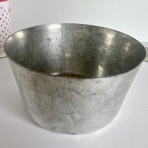 May include: A vintage, brushed metal bowl with a wide, curved rim and two small, petal-shaped handles. The bowl has a slightly tapered shape and a weathered appearance. A red and white polka-dotted container is partially visible in the background.