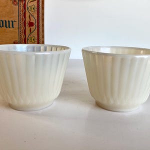Op de afbeelding: Twee crèmekleurige, geribbelde theekopjes met oren. De kopjes hebben een parelmoerglans en staan tegen een witte achtergrond. Een houten kist met het woord "Flour" is gedeeltelijk zichtbaar op de achtergrond.