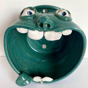 May include: A teal ceramic face-shaped bowl with a wide open mouth and large white teeth. The bowl has two eyes with blue irises and a small nose. A small leaf-like shape is on the bottom left. The bowl has a hole at the top for hanging.