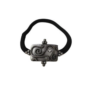 May include: A black hair tie with a rectangular, silver-colored embellishment. The embellishment features a rope-like design, small decorative spheres, and a single clear rhinestone. The hair accessory is set against a white background.