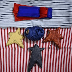 May include: A selection of craft materials in patriotic colours. Features red and blue ribbons, a ball of blue raffia, and star-shaped ornaments in yellow, red, and black. The items are arranged on striped fabric.