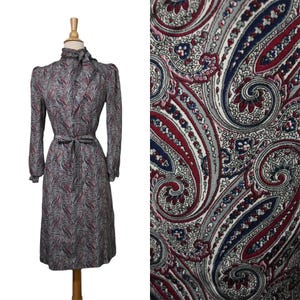 May include: A long-sleeved dress featuring a high neck with a tie detail and a waist belt. The garment is covered in a detailed paisley pattern consisting of red, navy blue, and white swirls on a light gray background.