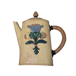 Pode incluir: Bule de cerâmica creme com um design floral. O bule apresenta uma flor azul com detalhes em rosa e verde, uma alça marrom e uma tampa correspondente. O bico e a alça são marrons. O bule é um item decorativo de cozinha.