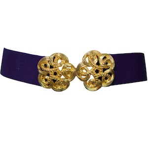 Puede incluir: Un cinturón de terciopelo morado con cierres dorados ornamentados. Los cierres tienen un diseño floral. El cinturón es de estilo gargantilla y está sobre un fondo blanco.
