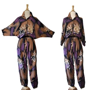 Puede incluir: Un conjunto de dos piezas que consta de una camisa abotonada con mangas anchas y pantalones a juego. La tela presenta un patrón abstracto en tonos marrón, morado y negro con detalles en forma de cuadrícula blanca en toda la prenda.