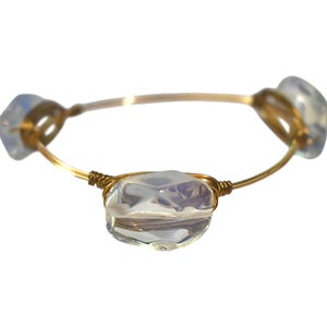 Peut inclure: Un bracelet en fil métallique doré orné de trois perles translucides, irisées et de forme irrégulière. Le fil est enroulé solidement autour de chaque perle, formant un anneau circulaire continu qui maintient les pierres sur la fine structure.