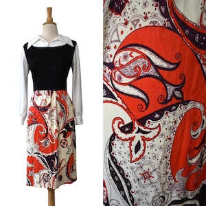 May include: A long-sleeve dress featuring a white pointed collar, a solid black bodice, and a skirt with a vibrant orange, dark purple, and white paisley pattern. The dress has a fitted waist and reaches below the knee, showcasing a detailed print.
