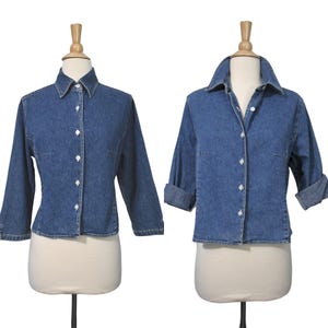 May include: Two denim shirts on mannequins. The shirts are a medium blue wash with white buttons and a collared neckline. One shirt has long sleeves, and the other has rolled-up sleeves. Both shirts have a classic, casual style.