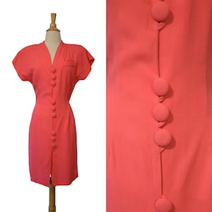 May include: Coral-colored dress with short sleeves and a V-neck. The dress features a row of large, matching buttons down the front and a small pocket detail. The dress is displayed on a mannequin.
