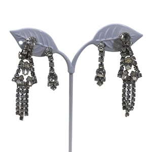 May include: A pair of silver-toned, dangling earrings with clear, sparkling rhinestones. One earring features a triangular design with multiple rhinestone strands, while the other has a simpler, elongated shape. The earrings are displayed on a white, leaf-shaped earring stand.