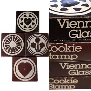 May include: A collection of vintage cookie stamps with various designs, including hearts, flowers, and a sunburst pattern. The stamps are arranged in a cross shape, with a box labeled "Vienna Glass Cookie Stamp" visible in the background.