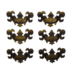 May include: Six antique brass drawer pulls with a decorative, ornate design. Each pull features a curved handle and a detailed backplate with floral-like accents. The pulls are arranged against a white background.