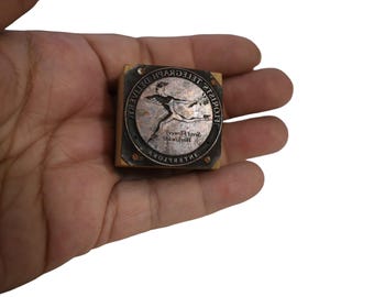Puede incluir: Un sello de metal vintage con una figura corriendo y las palabras "Florists Telegraph Delivery International". El sello es cuadrado, con una base de madera y una placa de metal con el diseño y el texto.
