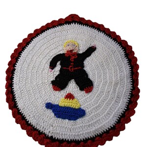 Puede incluir: Un salvamanteles circular hecho a mano, tejido a ganchillo, con un centro blanco, borde negro y rojo, y un diseño caprichoso. El diseño presenta una figura con un atuendo negro con detalles rojos y una tetera azul con detalles amarillos y rojos.