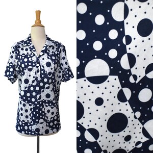 May include: A short-sleeved shirt featuring a navy blue and white geometric pattern of varying sized polka dots arranged in a patchwork style. The shirt has a collared neckline with a partial button-down front placket and a straight hemline.
