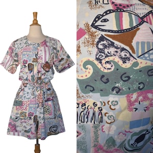 Könnte beinhalten: Ein Vintage-Romper mit einem farbenfrohen, abstrakten Muster. Der Romper hat kurze Ärmel, eine Knopfleiste vorne und eine Kordel in der Taille. Der Stoff ist hellblau mit rosa, braunen und grünen Formen, darunter Fische und andere Designs. Der Romper wird auf einer Schaufensterpuppe präsentiert.