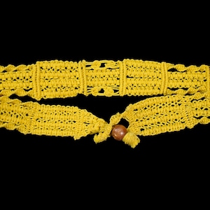 May include: A bright yellow macrame belt with a decorative pattern. The belt features a wooden bead at the end of the tie closure. The belt is shown against a black background.